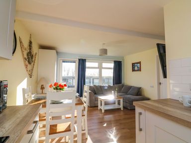 A dining area with a wooden table and white chairs next to a living room with a grey sofa and coffee table at The Sail Loft in Norton Park near Dartmouth