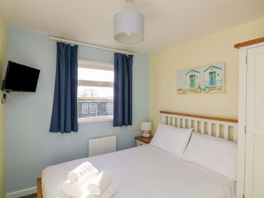 A bedroom with a bed with white linens and towels a wardrobe a nightstand a lamp a wall-mounted tv and a window with dark curtains at The Sail Loft in Norton Park near Dartmouth