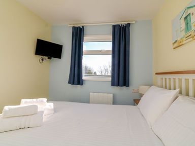 A bedroom with a double bed white towels on bed a wall-mounted tv and window with blue curtains at The Sail Loft in Norton Park near Dartmouth