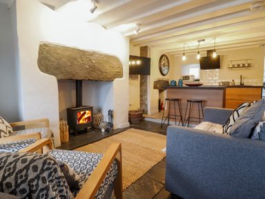 A living room with a wood burning stove two armchairs a gray sofa and a kitchen area with stools and hanging lights at Ty Dyffryn in Llan Ffestiniog