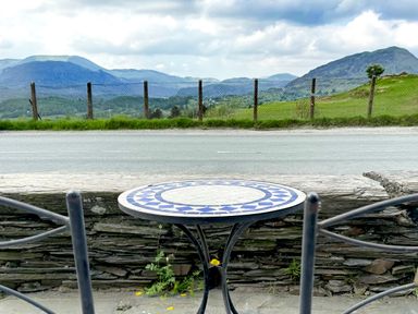 A round table with a blue and white tile top and two metal chairs facing a road with mountains in the background at Ty Dyffryn in Llan Ffestiniog