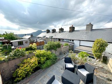 An outdoor patio with black wicker chairs and a table surrounded by plants and stone walls with houses and mountains in the background at Ty Dyffryn in Llan Ffestiniog