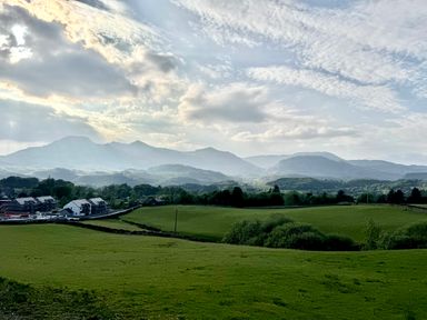 Green fields with scattered houses and mountains in the background under a cloudy sky at Ty Dyffryn in Llan Ffestiniog