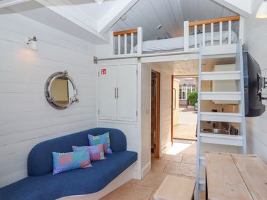 A small living area with a blue sofa and colorful cushions a wooden table with stools a loft bed accessed by a white ladder and a round porthole mirror on the wall at Beach Hut 5 in Shaldon