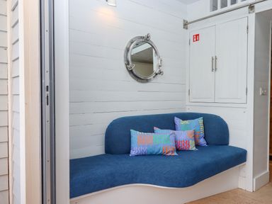 A corner seating area with a blue upholstered bench and colorful pillows under a round mirror and next to white cupboards at Beach Hut 5 in Shaldon
