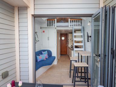 A compact living space with a blue sofa, loft bed, kitchen area, wooden door, and bar stools at Beach Hut 5 in Shaldon