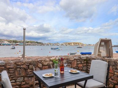 An outdoor patio table with plates of scones glasses and a bottle of wine overlooking a harbor with boats at Beach Hut 5 in Shaldon