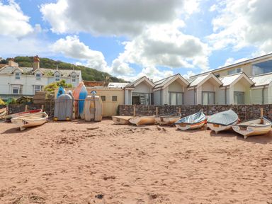 A sandy beach area with small boats lined up and stacked against a stone wall with houses in the background at Beach Hut 5 in Shaldon