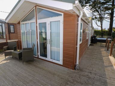 An outdoor wooden deck with two chairs and a table next to a brown-sided building with glass doors and windows at Gentle Tide L46 Hasguard Cross near Little Haven