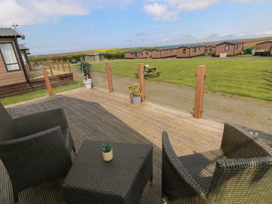 A wooden deck with outdoor wicker chairs and table overlooking a grassy area with several brown cabins at Gentle Tide L46 Hasguard Cross near Little Haven