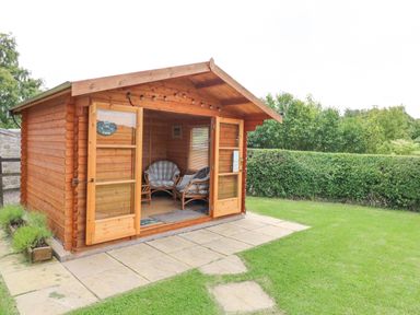 A wooden garden cabin with glass doors open showing wicker chairs inside at Loanview in Cornhill-On-Tweed