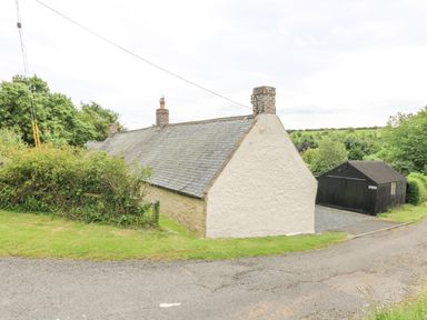 A rural cottage with a slate roof and two chimneys next to a black wooden garage surrounded by greenery at Loanview in Cornhill-On-Tweed