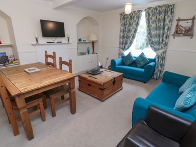 A living room with wooden dining table and chairs blue sofas a wooden chest and a wall mounted tv at White Swan Cottage in Alnwick