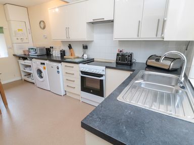 A kitchen with black countertops white cabinets a washing machine a small fridge an oven a toaster a microwave and a stainless steel sink at White Swan Cottage in Alnwick