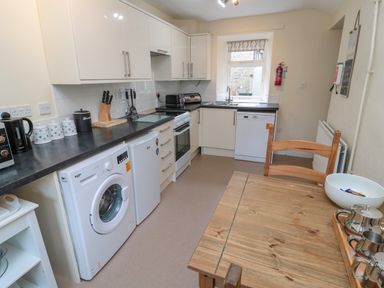 A kitchen with white cabinets a washing machine a fridge an oven and a wooden table with chairs at White Swan Cottage in Alnwick