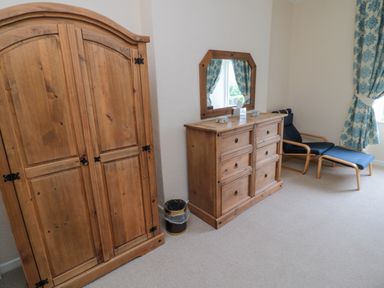 A room with a wooden wardrobe and dresser with mirror and a blue armchair with footrest at White Swan Cottage in Alnwick
