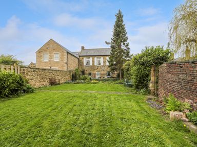 A backyard with green grass lawn stone and brick walls trees and a stone house at White Swan Cottage in Alnwick