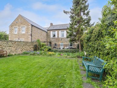 A garden with green grass benches and a stone house with white windows in the background at White Swan Cottage in Alnwick