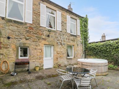 A stone house exterior with white door and windows a patio with a round glass table and four metal chairs a hot tub and a barbecue grill with a garden hose at White Swan Cottage in Alnwick