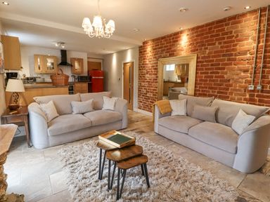 A living room with two beige sofas a wooden nesting table and a chandelier with an open kitchen and exposed brick wall at The Fullerton in Belsay