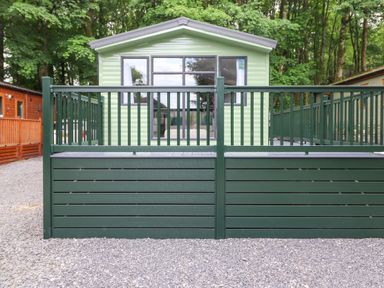 An outdoor view of a light green cabin with a dark green fenced deck surrounded by trees at Eltons Escape in Pony Meadow 3 White Cross Bay near Windermere