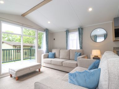 A living room with two beige sofas blue cushions a beige ottoman a round mirror and a window with blue curtains at Eltons Escape in Pony Meadow 3 White Cross Bay near Windermere