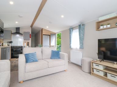 A living room with a beige sofa and blue cushions a television on a wooden stand and a kitchen area in the background at Elton's Escape in Pony Meadow 3 White Cross Bay near Windermere