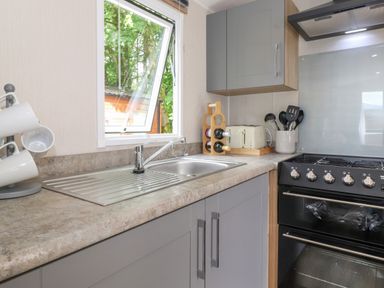 A kitchen countertop with a sink a rack of white mugs a wine rack a toaster cooking utensils and a black stove at Eltons Escape in Pony Meadow 3 White Cross Bay near Windermere