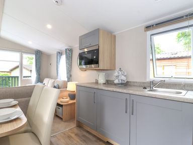 A kitchen area with gray cabinets a microwave kettle and cup holder next to a window and a dining area with white chairs and a table set with bowls next to a living room with a sofa and curtains at Eltons Escape in Pony Meadow 3 White Cross Bay near Windermere