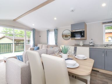 A dining table set with dishes and glasses next to a kitchen area with grey cabinets and a microwave and a living room with sofas and sliding glass doors at Eltons Escape in Pony Meadow 3 White Cross Bay near Windermere