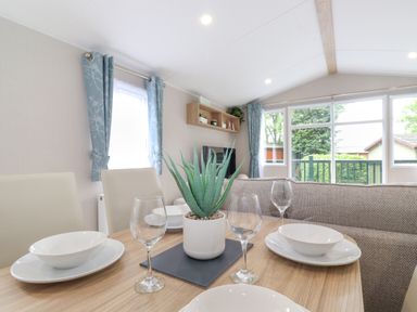 A dining area with table set for four and a plant in the center beside a living room with windows and curtains at Eltons Escape in Pony Meadow 3 White Cross Bay near Windermere