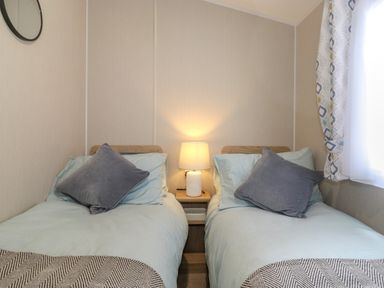 A bedroom with two single beds each with a pillow a bedside table with a lamp and a window with patterned curtains at Eltons Escape in Pony Meadow 3 White Cross Bay near Windermere