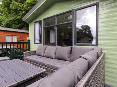 A patio with a gray cushioned wicker sofa and a wooden table outside a green cabin at Elton's Escape Pony Meadow 3 White Cross Bay near Windermere