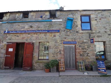 A stone building with signs for Swallow Fish Ltd Traditional Smokehouse and Fisherman's Kitchen with red doors and fishing equipment outside at Greenlee House in Seahouses