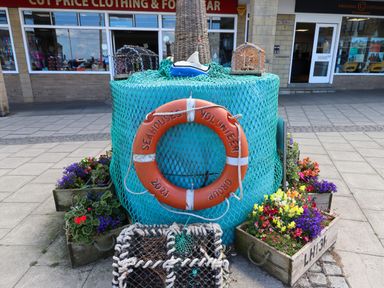 A fisherman's display with a life buoy, fishing nets, traps, and flower boxes on a pavement outside shops in Seahouses
