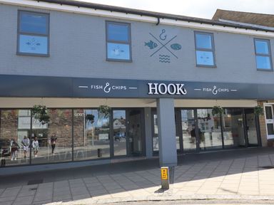 The exterior of a fish and chips shop named Hook with hanging flower baskets and glass windows displaying reflections of people and buildings