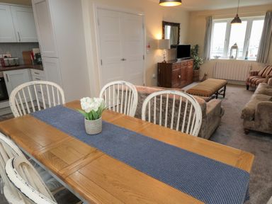 A dining table with white chairs and a flower vase in a living room with sofas a TV and window at Greenlee House in Seahouses