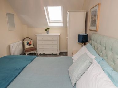 A bedroom with a bed blue bedding a white chest of drawers a chair with rooster pillow a wardrobe and a window at Greenlee House in Seahouses