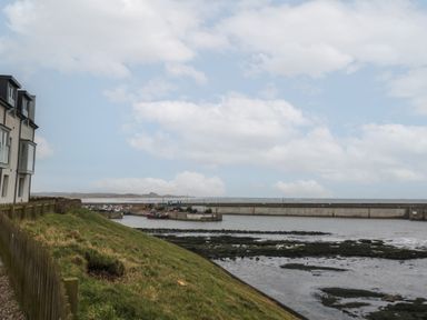 A waterfront view with a grassy slope a wooden fence a building on the left and a harbor with boats and a breakwater at Greenlee House in Seahouses