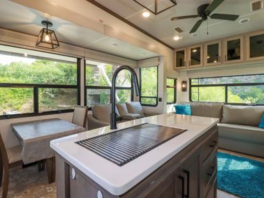 An interior view of an RV kitchen and living area with a kitchen island featuring a sink and faucet a dining table with chairs beige sofas with blue pillows and large windows at Tickeydoo Tor Down Quarry in St Breward