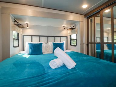A bedroom with a teal bedspread and pillows two rolled white towels on the bed bedside windows and a mirrored wardrobe at Tickeydoo Tor Down Quarry in St Breward