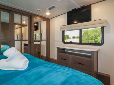 A bedroom with a bed covered in blue bedding two rolled white towels on the bed a mirrored closet a dresser with two drawers a window with a view of greenery and a wall-mounted television
