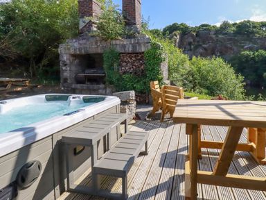 An outdoor deck area with a hot tub wooden benches a stone fireplace and trees in the background at Tickeydoo Tor Down Quarry St Breward