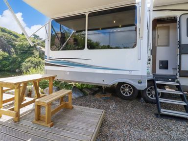 An outdoor wooden picnic table and bench set next to a white caravan with stairs on a gravel surface at Tickeydoo Tor Down Quarry in St Breward