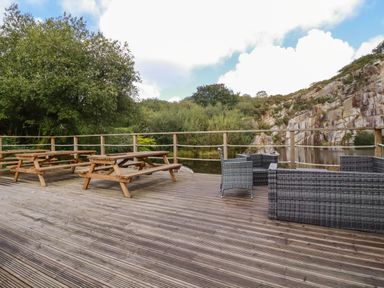 An outdoor wooden deck with picnic tables and wicker furniture overlooking a pond and rocky hillside at Tickeydoo Tor Down Quarry in St Breward