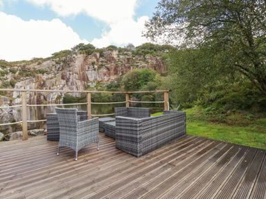 An outdoor wooden deck with gray wicker chairs and a table overlooking a rocky lake with trees at Tickeydoo Tor Down Quarry in St Breward