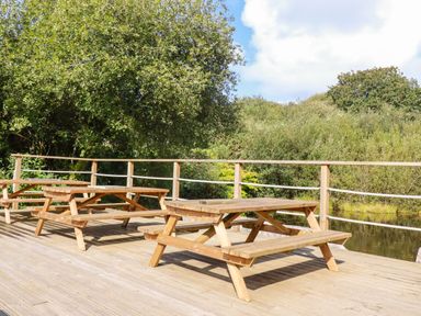 An outdoor wooden deck with picnic tables and railing by trees and a small body of water at Tickeydoo Tor Down Quarry in St Breward