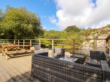 An outdoor wooden deck with wicker seating and a picnic table overlooking water and rocky hillside at Tickeydoo Tor Down Quarry in St Breward