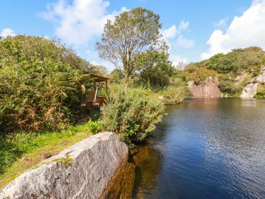A lake with surrounding trees bushes and rocks with a small wooden shelter at Tickeydoo Tor Down Quarry in St Breward