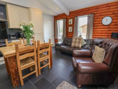 A dining area with wooden table and chairs next to a sitting area with leather sofas and log cabin walls at The Blacktail in Llanerch Park near St Asaph